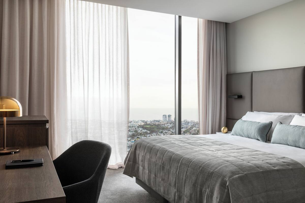 A hotel room with a large bed, a desk, and a view of a cityscape through large windows.