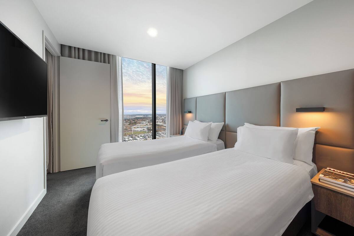 A hotel room with two beds, a large window with a city view, a flat-screen TV mounted on the wall, and a bedside table with a lamp.