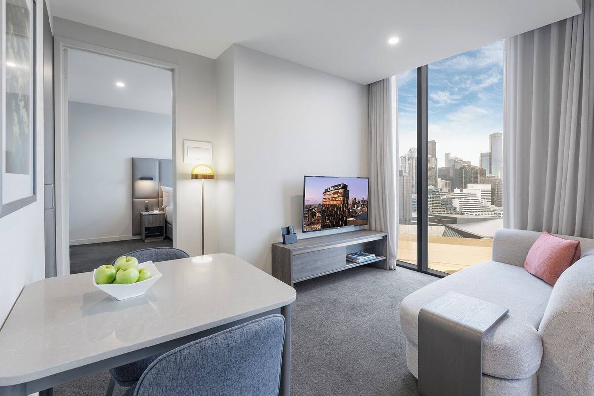 A modern hotel room with a dining area, a sofa, a TV, and a city view.