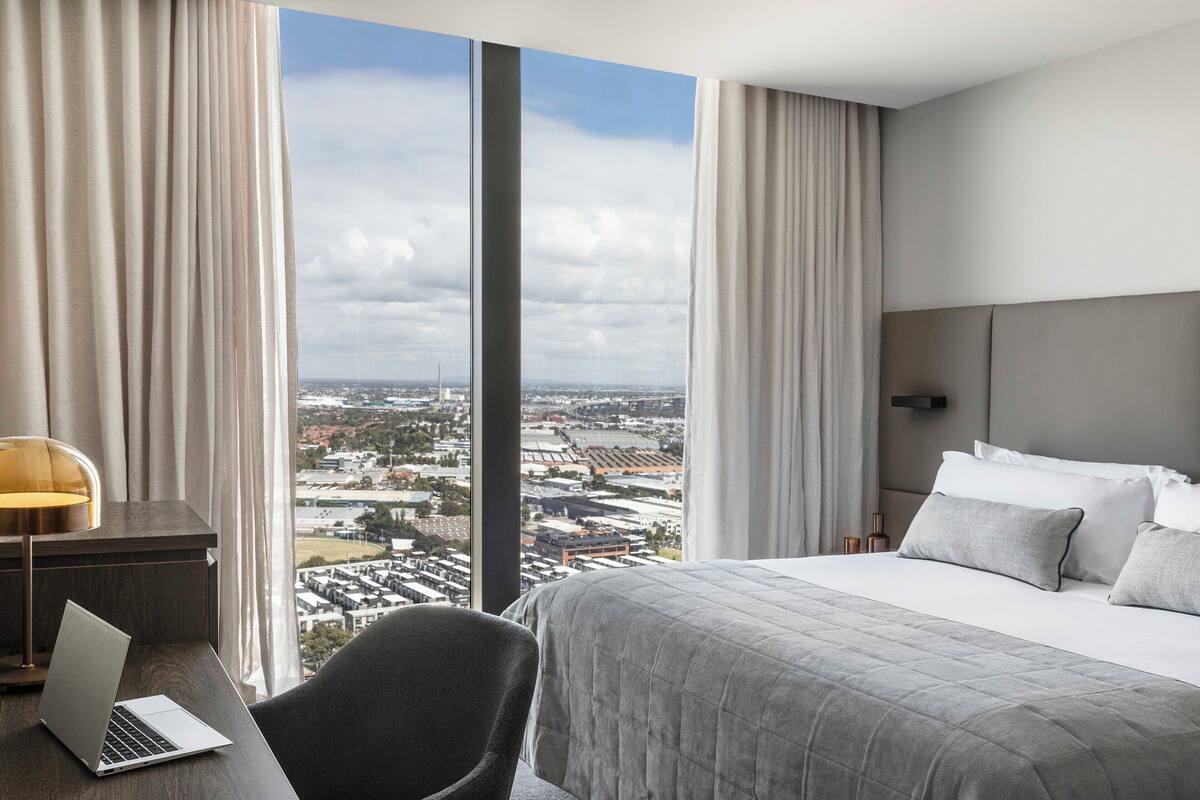 A hotel room with a large bed, a desk with a laptop, and a view of a cityscape through large windows.