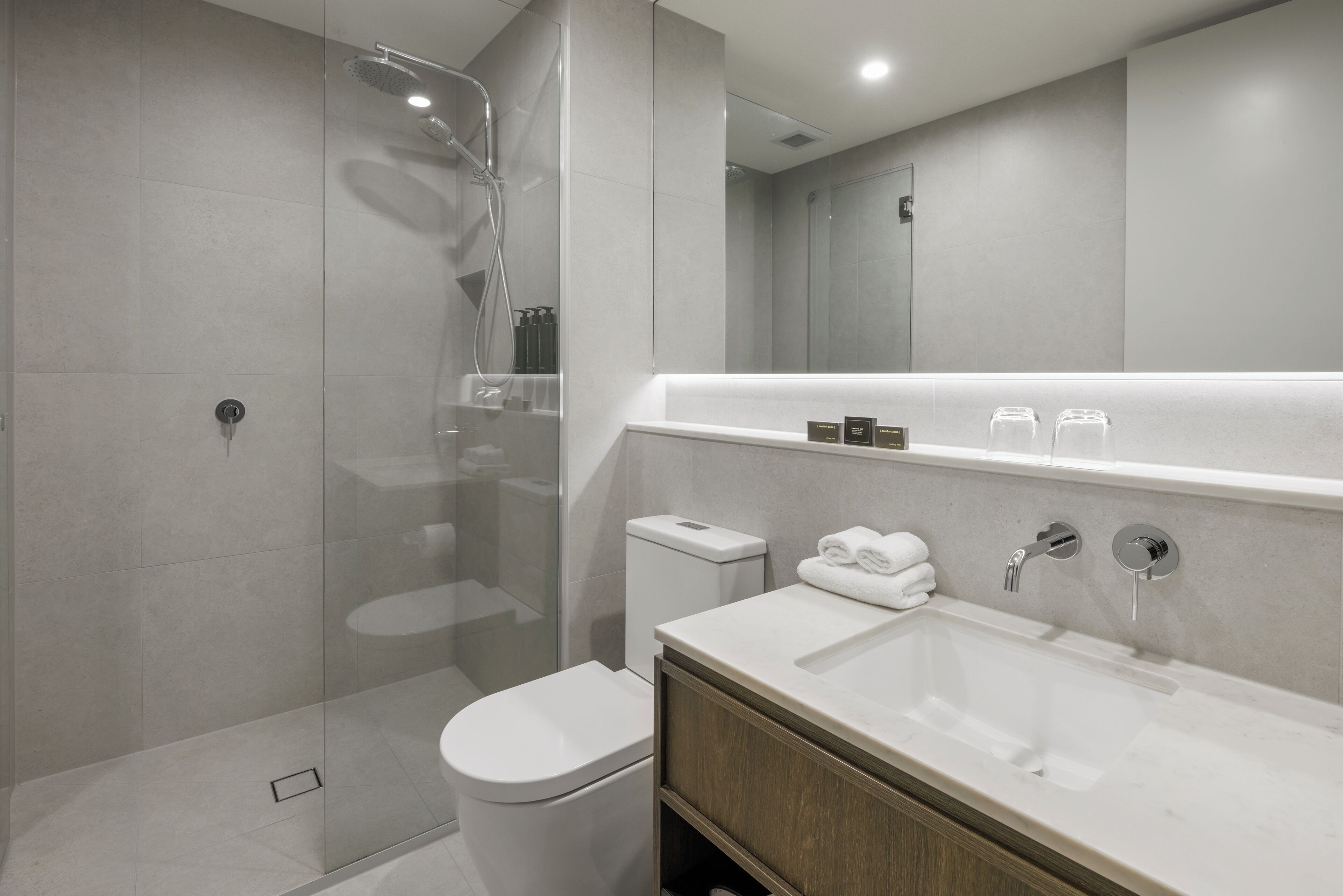 A modern bathroom with a glass-enclosed shower, a white toilet, a wooden vanity with a sink, and a large mirror above the vanity.
