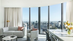 Ein modernes Hotelzimmer mit Blick auf die Stadt, inklusive Sofa, Sessel, kleinem Tisch und einem Essbereich mit Blumenvase.