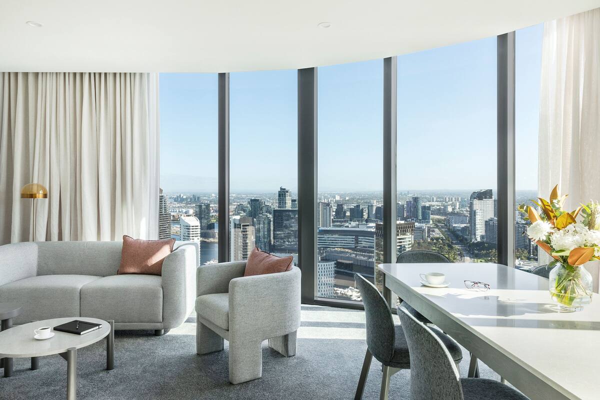 A modern hotel room with a city view, a sofa, a chair, a small table, and a dining area with a vase of flowers.