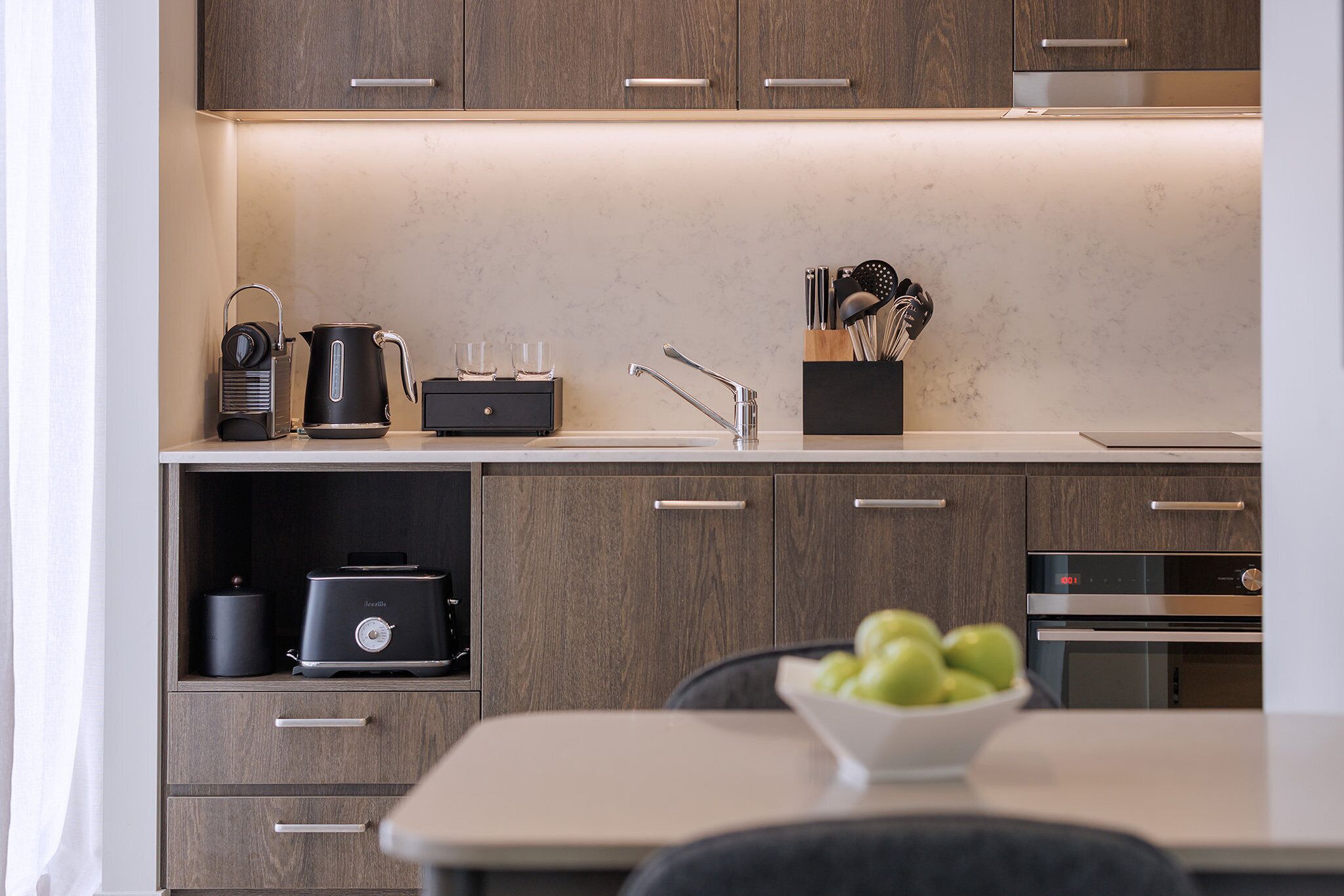 A modern kitchen with wooden cabinets, a countertop, a kettle, a toaster, and a bowl of apples.