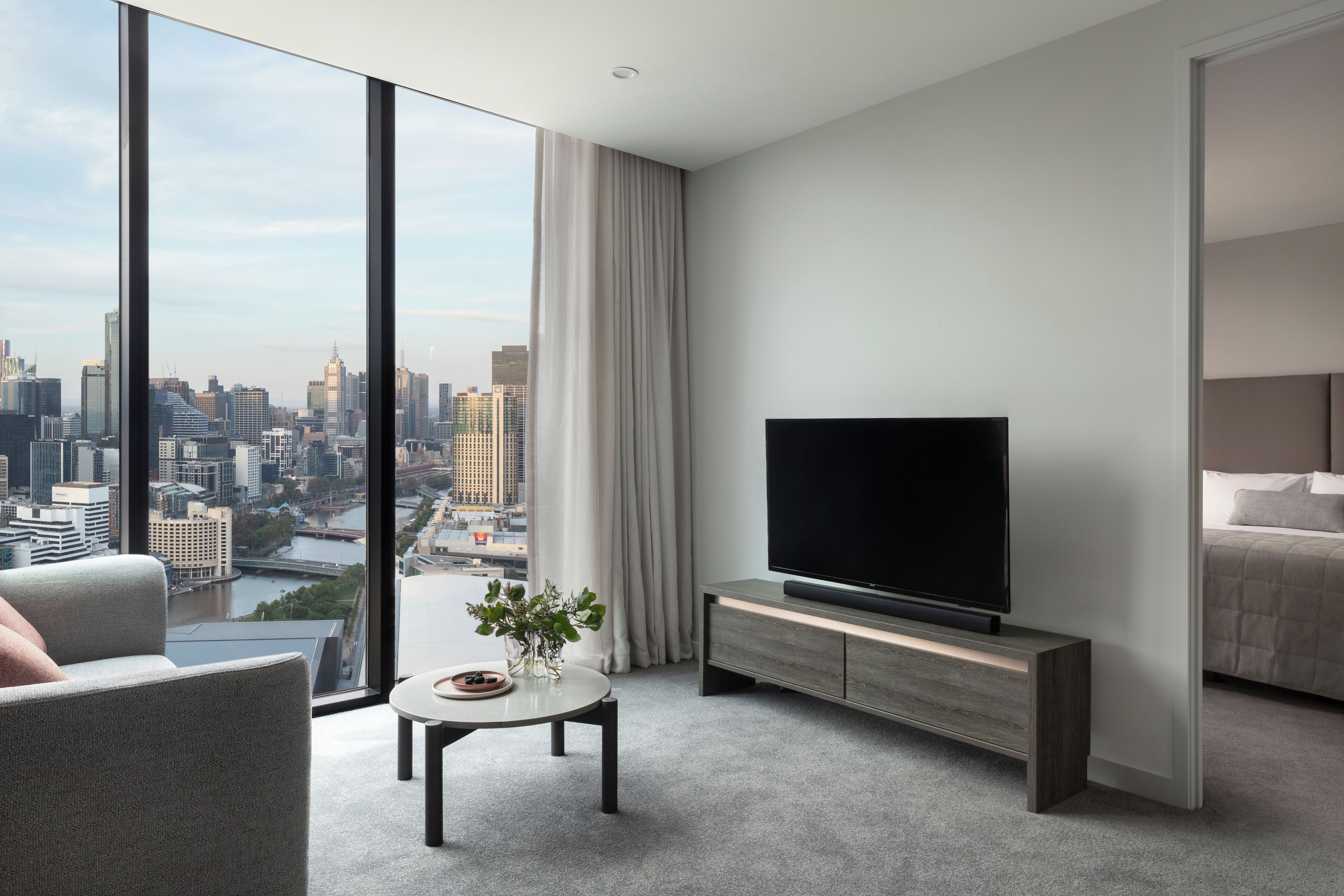 Habitación de hotel moderna con un ventanal que ofrece vistas a la ciudad, un televisor de pantalla plana sobre un mueble de madera, una pequeña mesa redonda con un florero y un área de descanso confortable.