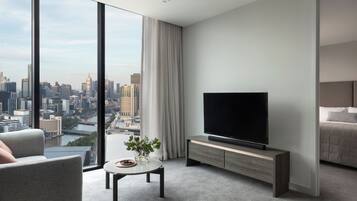 Habitación de hotel moderna con un ventanal que ofrece vistas a la ciudad, un televisor de pantalla plana sobre un mueble de madera, una pequeña mesa redonda con un florero y un área de descanso confortable.