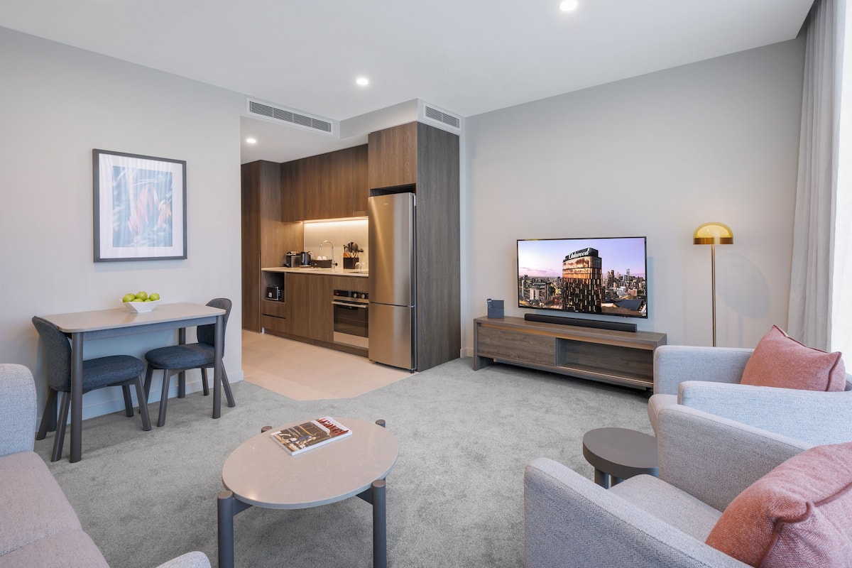A modern living room with a sofa, a coffee table, a TV, and a kitchen area with a refrigerator and a dining table.
