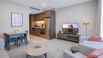 A modern living room with a sofa, a coffee table, a TV, and a kitchen area with a refrigerator and a dining table.