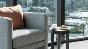 Ein modernes Zimmer mit einer grauen Couch, einem kleinen runden Tisch mit Speisen und einer Tasse, und einem großen Fenster mit Blick auf die Stadt.
