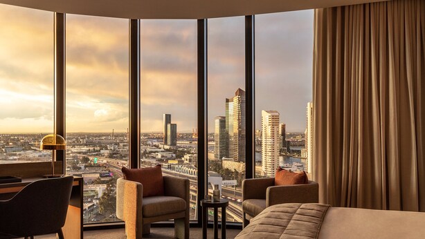 Ein Hotelzimmer mit großem Fenster und Blick auf die Stadt, einem Schreibtisch mit Lampe sowie zwei Sesseln.
