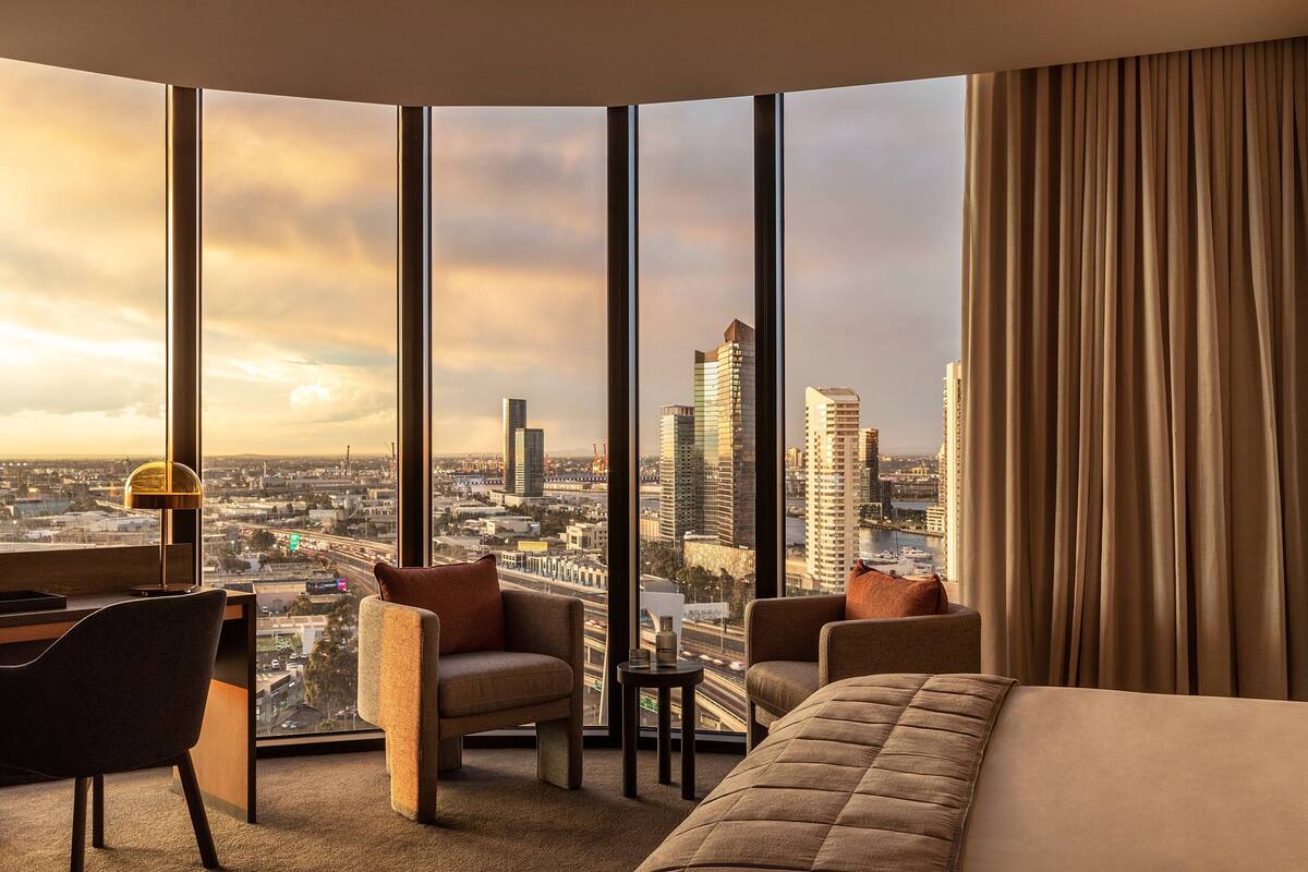 A hotel room with a large window offering a city view, a desk with a lamp, and two armchairs.