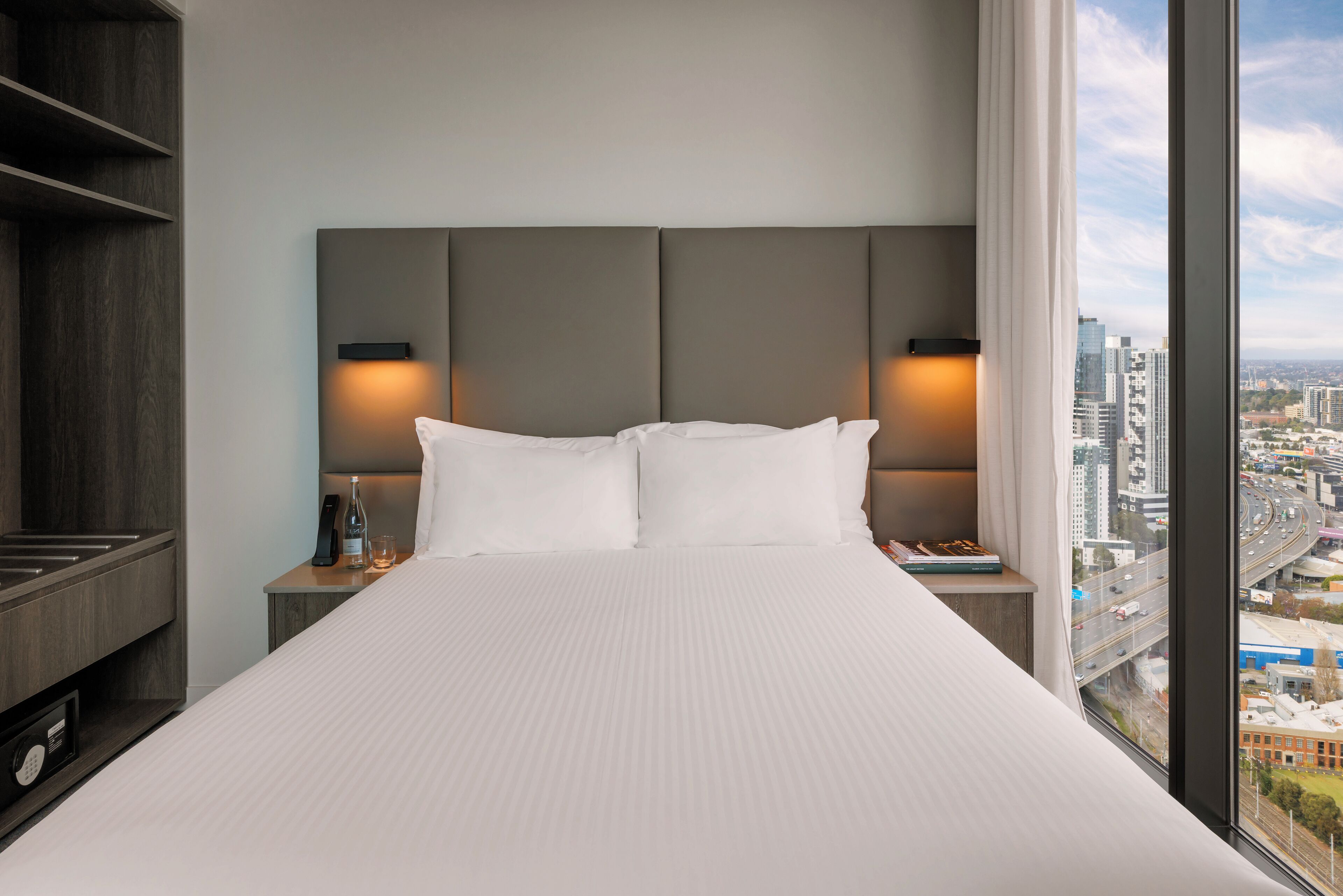 A hotel room with a large bed, two bedside tables, a nightstand, a lamp, and a view of the city through a large window.
