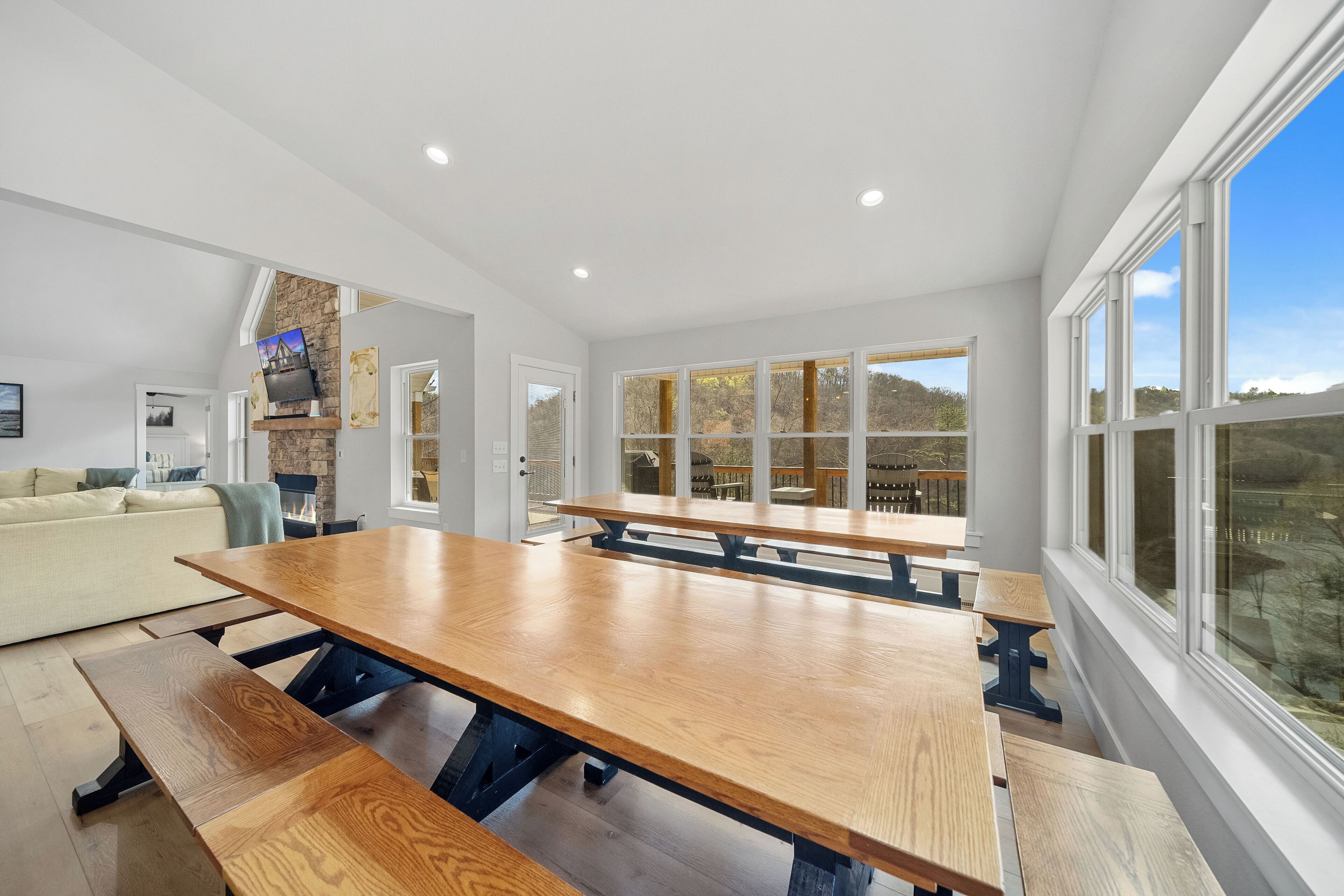 A spacious dining area with a large wooden table and benches, a stone fireplace, and large windows offering a view of the outdoors.