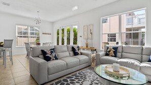A spacious living room with a grey sofa, a glass coffee table, and a dining area with a white table and chairs. - Private Pool | Reunion Membership | 8 Miles to Disney (Kissimmee)