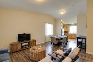 A spacious living room with a flat-screen TV, a wicker ottoman, and a sofa with cushions. A dining area with a table and chairs is visible in the background.