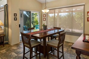Uma área de refeições com mesa de madeira, quatro cadeiras, luminária pendente e vista para um pátio externo por meio de uma porta de vidro deslizante.