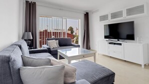 A modern living room with a grey sofa, a coffee table, a TV on a white cabinet, and a view of buildings through large windows. - Apartment Sleeps 6 with Pool (Fuengirola)