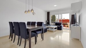 A modern living room with a dining area, featuring a sofa, a coffee table, and a dining table with chairs. There is a TV on a stand and a window with curtains. - Apartment Sleeps 6 with Pool (Fuengirola)
