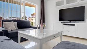 A modern living room with a large flat-screen TV, a grey sofa, a white coffee table, and a large window with a view of buildings. - Apartment Sleeps 6 with Pool (Fuengirola)