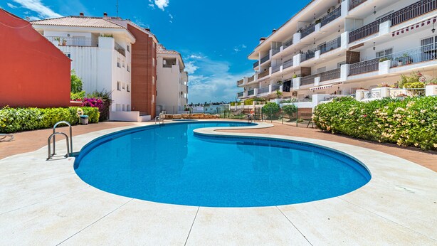 A swimming pool surrounded by multi-story buildings with balconies, a clear blue sky, and well-maintained landscaping. - Apartment Sleeps 6 with Pool (Fuengirola)