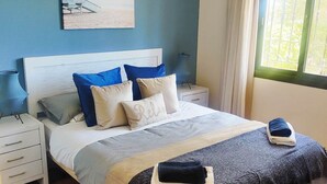 A bedroom with a bed, nightstands, a ceiling fan, a window with curtains, and a beach-themed painting on the wall.