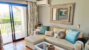 A living room with a beige sofa, a glass coffee table, and a view of a garden through a sliding glass door.
