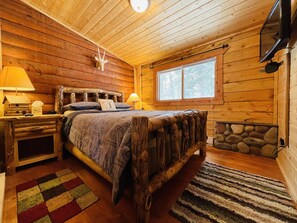 A cozy log cabin bedroom with a bed, two bedside lamps, a window, and a stone wall.