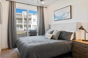 Ett sovrum med en stor säng, grå gardiner, utsikt över en byggnad utanför och en inramad tavla på väggen.