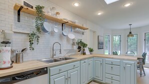 A modern kitchen with white brick backsplash, light blue cabinets, and a wooden countertop. There are shelves with dishes and plants, and a dishwasher is visible.