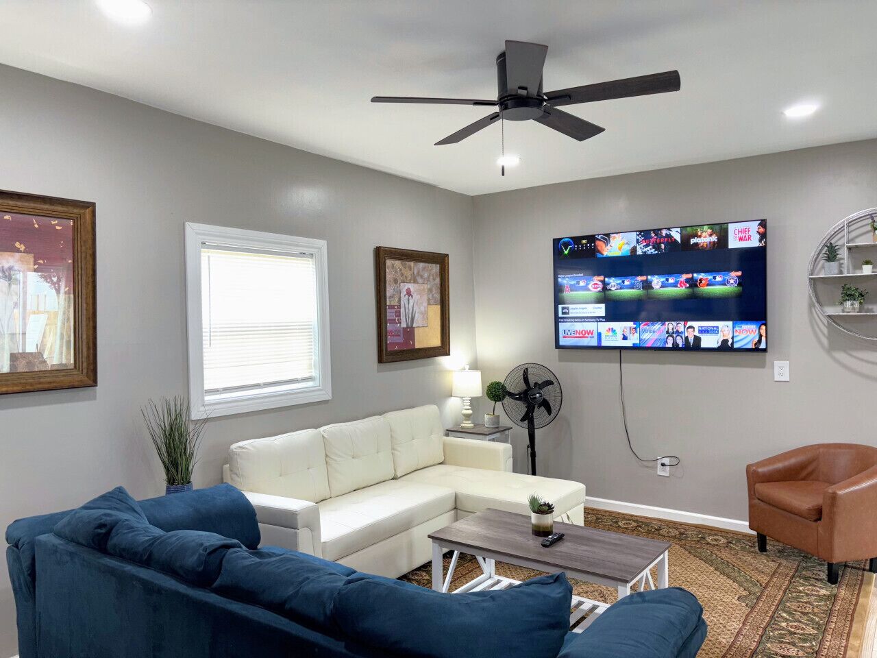 Una sala de estar con un sofá blanco tipo sectional, un sofá azul, una mesa de centro, un ventilador de techo, un televisor mostrando varios canales y una planta pequeña.