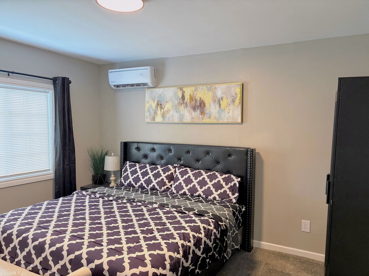 A bedroom with a tufted headboard, a large bed with patterned bedding, a wall-mounted air conditioner, and a painting above the bed.