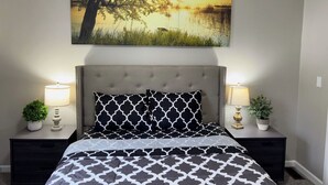 A bedroom with a bed, two nightstands, a painting of a tree and water, and two lamps.