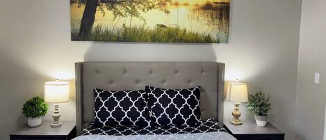 A bedroom with a bed, two nightstands, a painting of a tree and water, and two lamps.