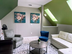 Uma sala de estar moderna com esquema de cores cinza e branco, apresentando uma poltrona cinza, um sofá azul, um sofá branco e uma mesa de centro redonda com uma pequena planta. Há duas pinturas florais na parede e um clarabóia acima da área de estar.