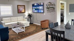 A living room with a white sofa, a coffee table, a TV mounted on the wall, a dining table with chairs, and a ceiling fan.