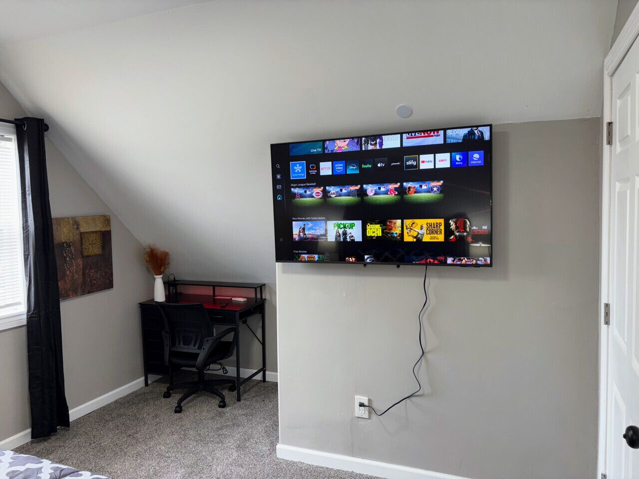 Een kamer met een tv aan de muur waarop verschillende streamingdiensten worden getoond, een bureau met een stoel en een schilderij.