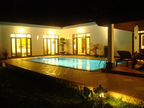 A swimming pool with a tiled deck, surrounded by walls with doors and windows, and potted plants.