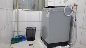Een kleine badkamer met een grijze wasmachine, een bezem en een prullenbak.