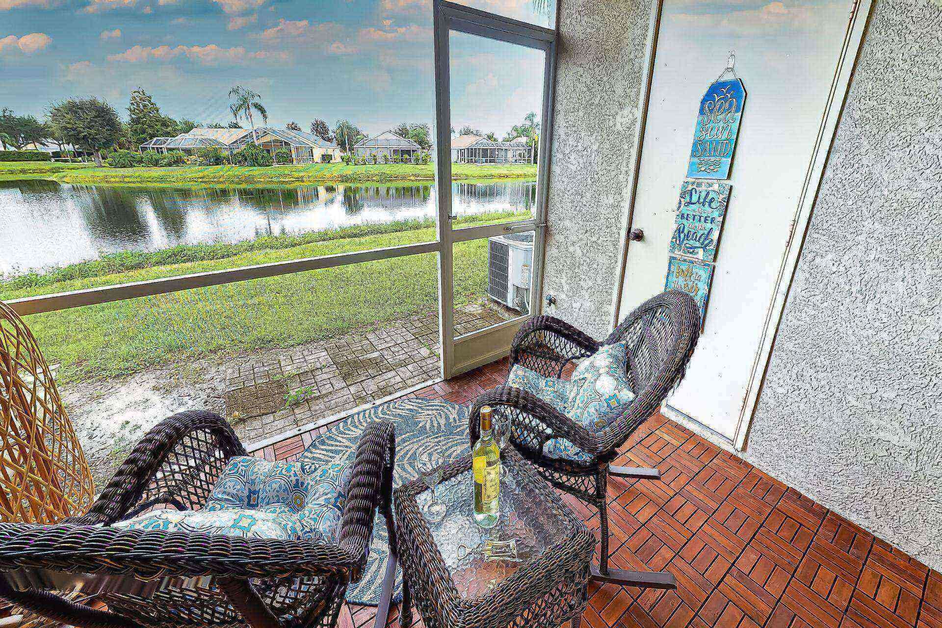 A room with a view of a water body and greenery, a wicker chair, a small table with a bottle, and a wall with a decorative sign.