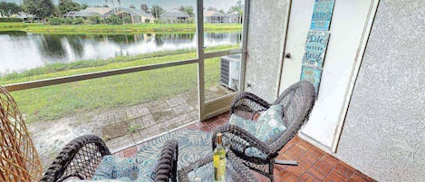 A room with a view of a water body and greenery, a wicker chair, a small table with a bottle, and a wall with a decorative sign.