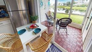 La cuisine est équipée d’un réfrigérateur en acier inoxydable, et on y trouve une table à manger avec de l’assiette bleue, ainsi que des chaises en osier. À l’extérieur, une porte vitrée donne accès à une terrasse d’où l’on peut admirer un étang et la verdure environnante.