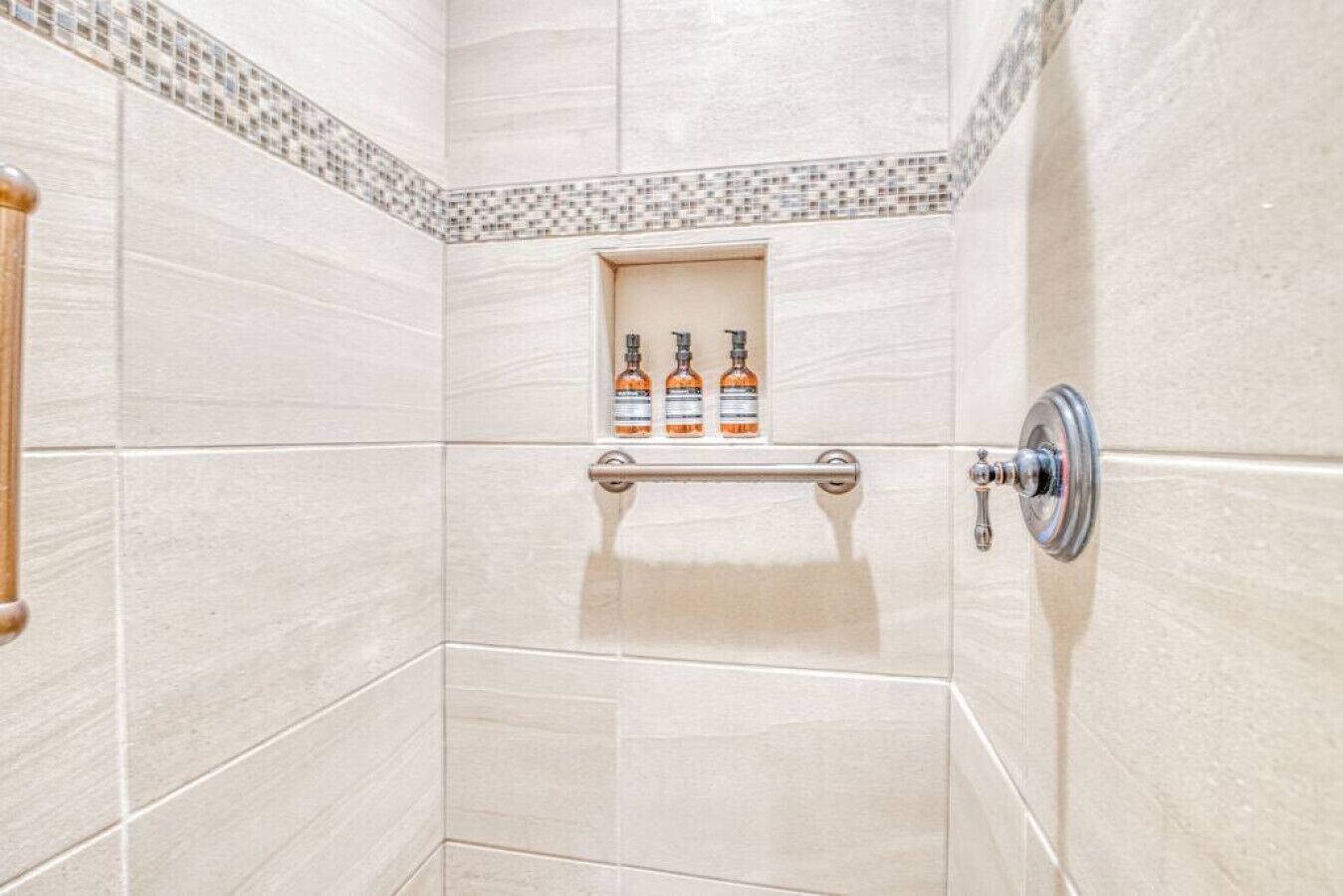 Un espace douche avec des carreaux de couleur beige, une étagère en verre où sont posées des bouteilles et un robinet de commande pour la douche.