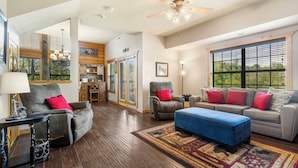 A spacious living room with a gray sofa, blue ottoman, and a gray armchair with red cushions. A wooden ceiling fan and large windows provide natural light. - Relaxing Walk-in Cabin in Branson Theatre District, Golf and Ticket Discounts (Branson)