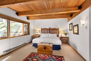 A bedroom with a wooden ceiling, a large bed, two bedside tables, a nightstand, a wooden chest, and a patterned rug.