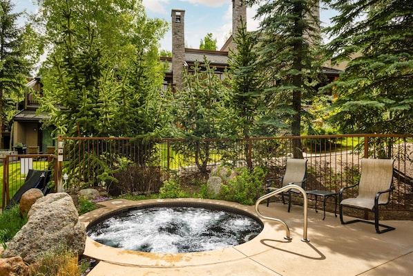 A hot tub surrounded by rocks and trees, with a wooden deck and chairs.