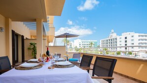A dining area with a table set for two, outdoor chairs, a table umbrella, and a view of buildings in the background. - Apartment Drac Blanc 1-4 close to the Beach with Sea View, Pool, Terrace & Wi-Fi (CALA RATJADA)