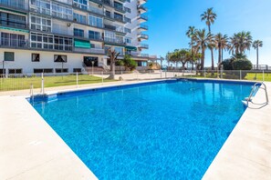A swimming pool with a clear blue water, surrounded by a white building with balconies, palm trees, and a fenced area. - Sunny Seaside Studio with Balcony, A/C & Sea View (Benalmádena Costa)