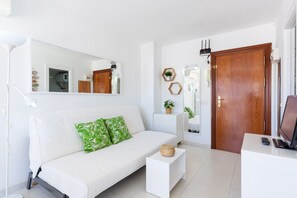 Un sofá blanco con almohadas verdes, una mesita auxiliar blanca, una puerta de madera, una consola blanca con un televisor y un espejo grande en la pared.