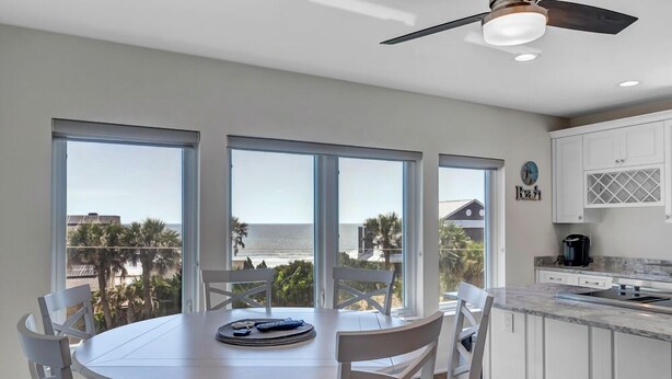 Un comedor con una mesa redonda y sillas, ventanas amplias con vistas a palmeras y edificios, y una cocina con gabinetes blancos y una encimera.