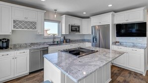 Una cocina moderna con armarios blancos, una isla central, electrodomésticos de acero inoxidable y un amplio ventanal que proporciona luz natural.